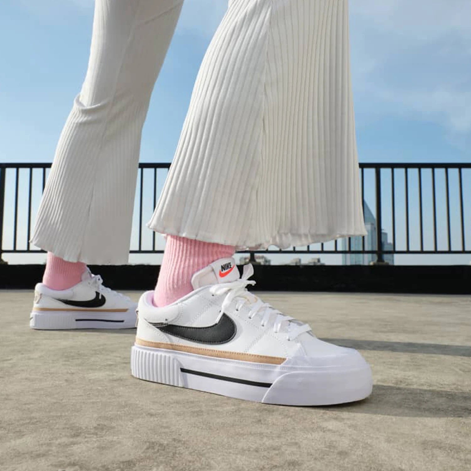 White sneakers with black swoosh logo worn by a person on a rooftop.